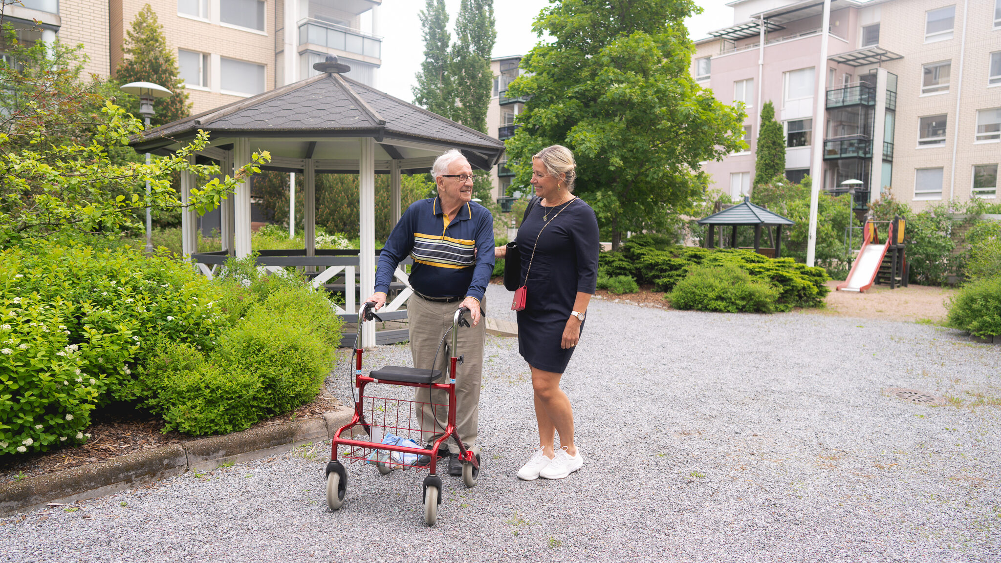 Iäkäs mies kävelee rollaattorin kanssa pihalla, nainen juttelee hänen kanssaan